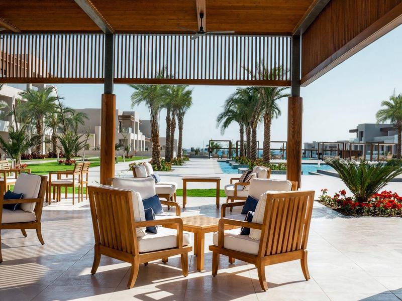 Espace extérieur sous un toit en bois avec des chaises et tables en bois face à des palmiers et une piscine.