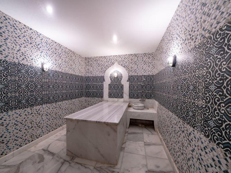 Ein heller Hammam-Raum mit marmoriertem Tisch, Wänden mit blauen und weißen Ornamenten und Bogenfenster.