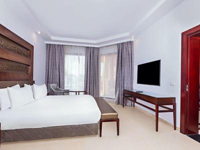 Lichte hotelkamer met groot bed, wanddecoratie, tv en balkondeuren met gordijnen.