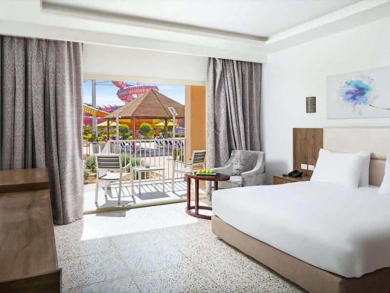 Lichte hotelkamer met balkon met uitzicht op zwembad en tropisch resort onder blauwe lucht.