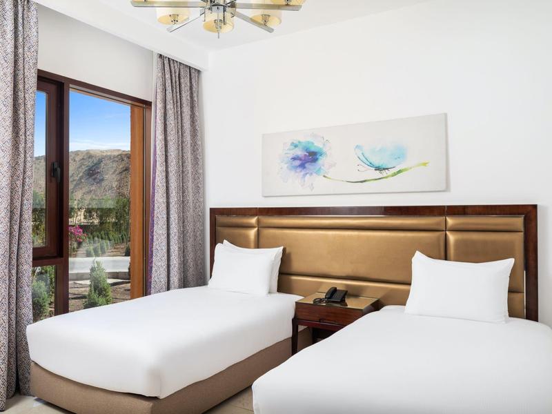 Habitación de hotel con dos camas individuales, mesa de noche y vista a montañas a través de una ventana.