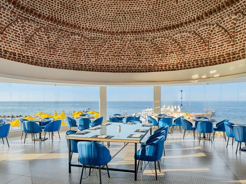 Restaurante elegante con sillas azules y vista al mar bajo una cúpula artística.