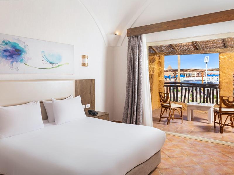 Habitación de hotel luminosa con cama blanca y balcón con vistas al mar y barcos.