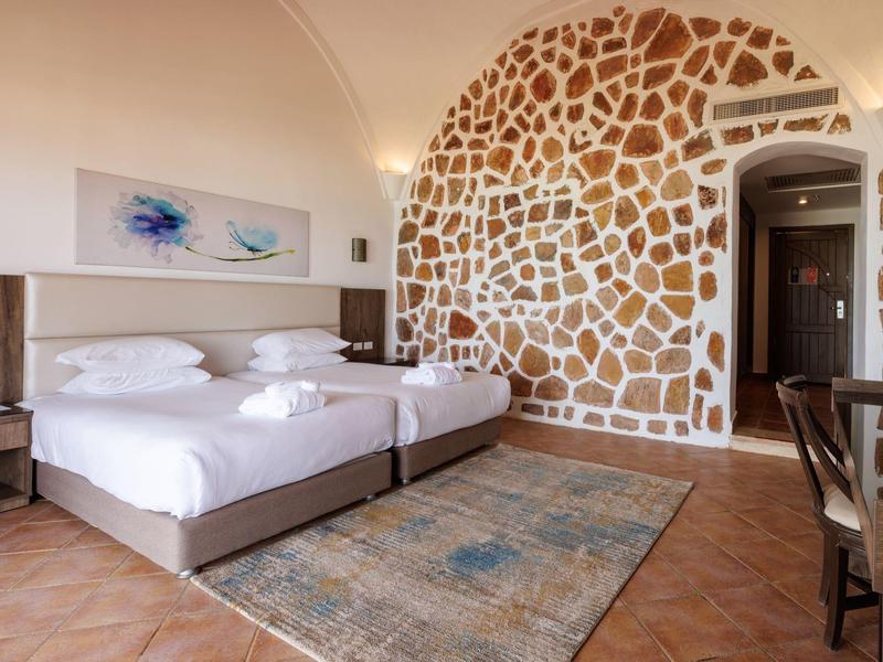 Habitación de hotel luminosa con cama doble, pared decorativa de piedra y alfombra sobre suelo de baldosas.