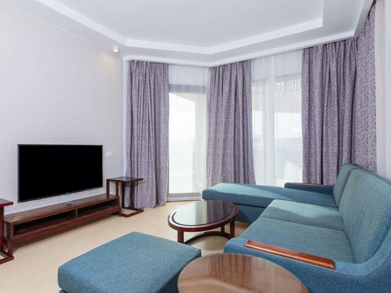 Soggiorno d'hotel moderno con divano ad angolo blu, TV e tende grigie.