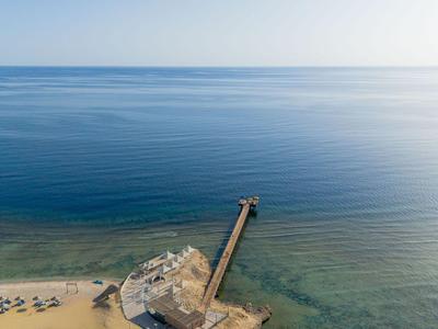 Aerial view of a long pier with a building at the end, surrounded by clear water and sandy beach with loungers.