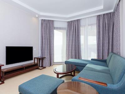 Soggiorno d'hotel moderno con divano ad angolo blu, TV e tende grigie.