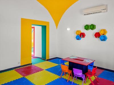 Kleurrijke kinderspeelruimte met tafels, stoelen en wandplanken in een witte kamer met gele accenten.