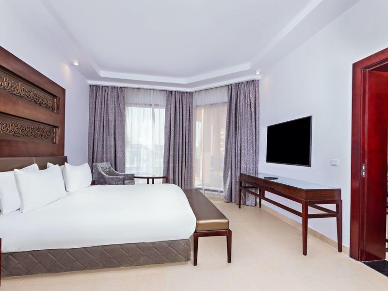 Camera d'hotel spaziosa con letto matrimoniale, TV a parete e grandi finestre con tende.