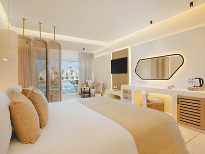 Chambre d'hôtel lumineuse avec grand lit, décor moderne, TV et vue sur balcon.