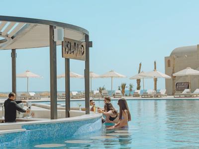 Piscina dell'hotel con bar in acqua, ospiti seduti che ordinano bevande, ombrelloni sullo sfondo.
