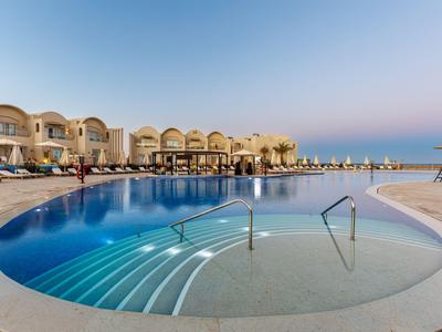 Großer runder Pool mit Stufen und Handläufen vor einem Hotel und dem Meer bei klarem Himmel.