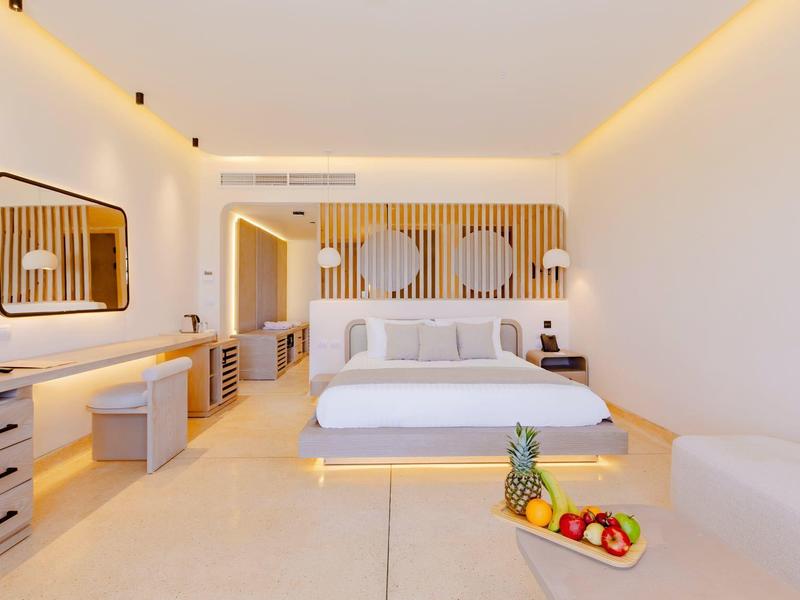 Habitación de hotel moderna con cama doble, área de descanso y cesta de frutas sobre suelos claros.