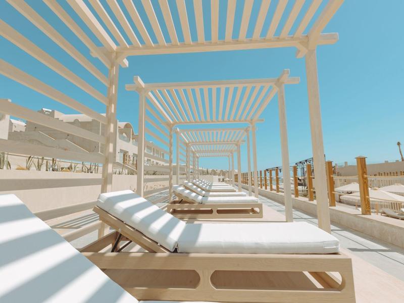 Sunbeds under white pergolas on a sandy beach with clear blue sky.