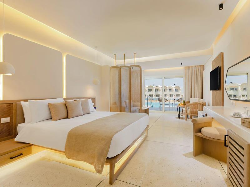 Bright hotel room with large bed, seating area, and balcony view of the sea.