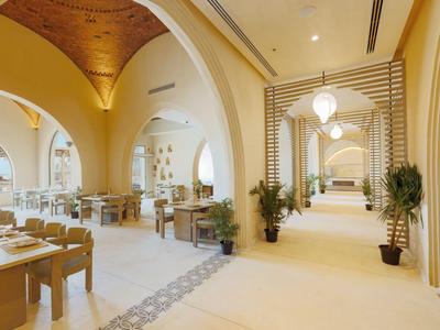 Jasna sala restauracyjna z łukowatymi oknami, drewnianymi stołami i roślinami wzdłuż korytarza.