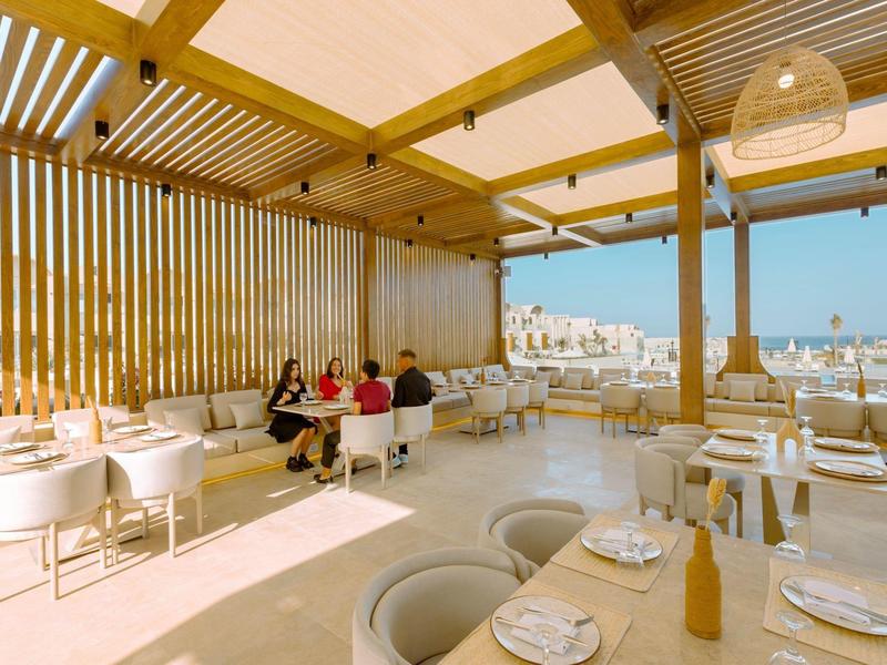 Terrasse extérieure avec tables rondes, chaises blanches et pergola en bois avec vue sur la mer.