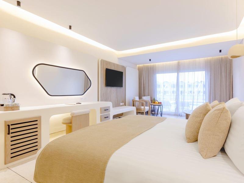 Chambre d'hôtel lumineuse avec grand lit, décoration moderne, TV et balcon avec rideaux.