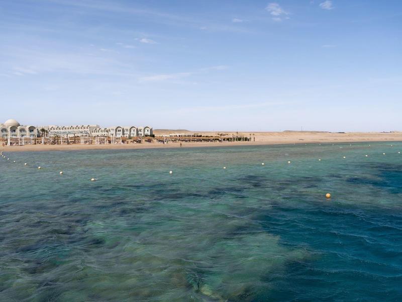 Clear blue sea water near a sandy beach with thatched umbrellas under a blue sky.