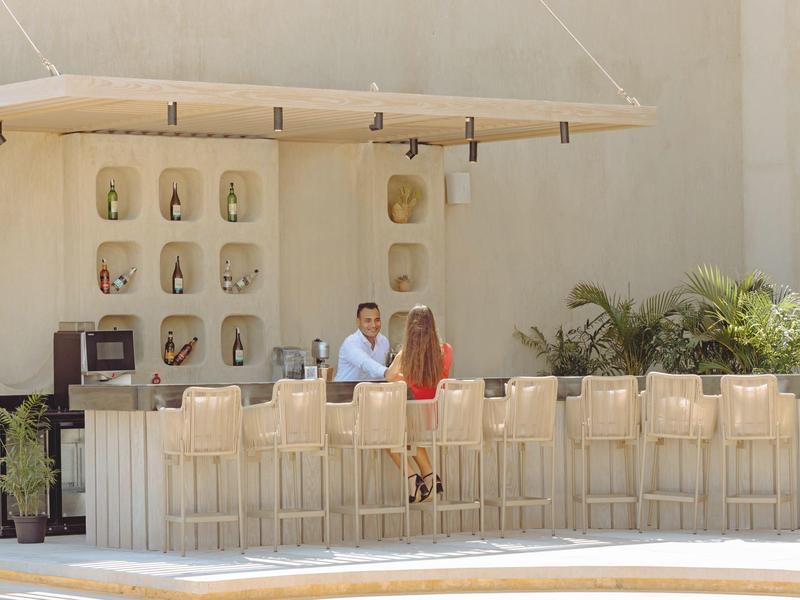 Outdoor poolside bar with two people chatting amid tall beige stools and potted plants.
