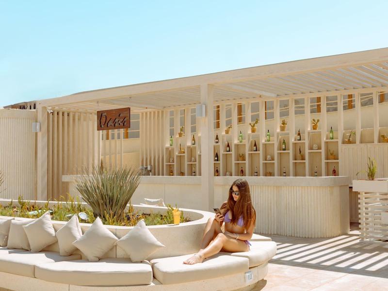 Woman sitting on white outdoor lounge chairs near a modern pool area with bottle display wall.