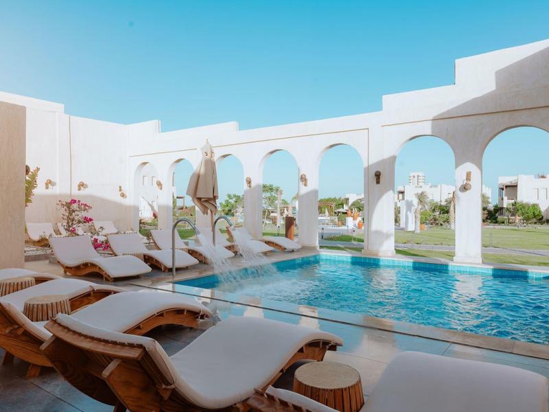 Lettini a bordo piscina sotto archi bianchi con cielo blu e prato verde sullo sfondo