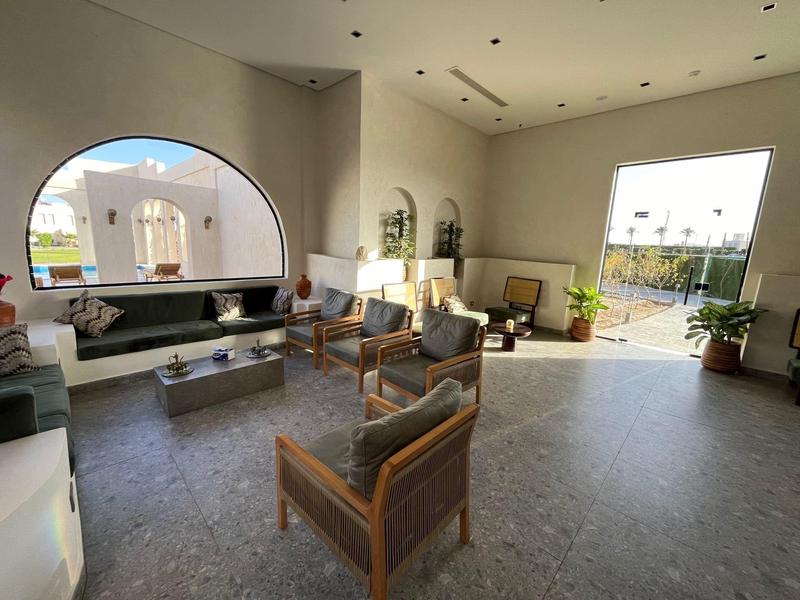 Heldere hotel lobby met moderne stoelen, planten, grote boogvormige raam en open deur naar buiten.