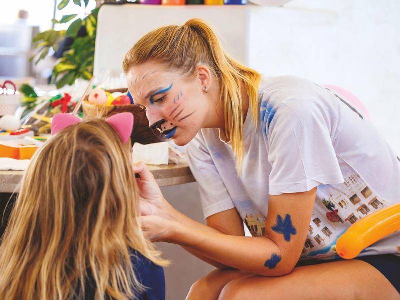 Une femme avec du maquillage peint applique du maquillage à une jeune fille dans une pièce lumineuse.