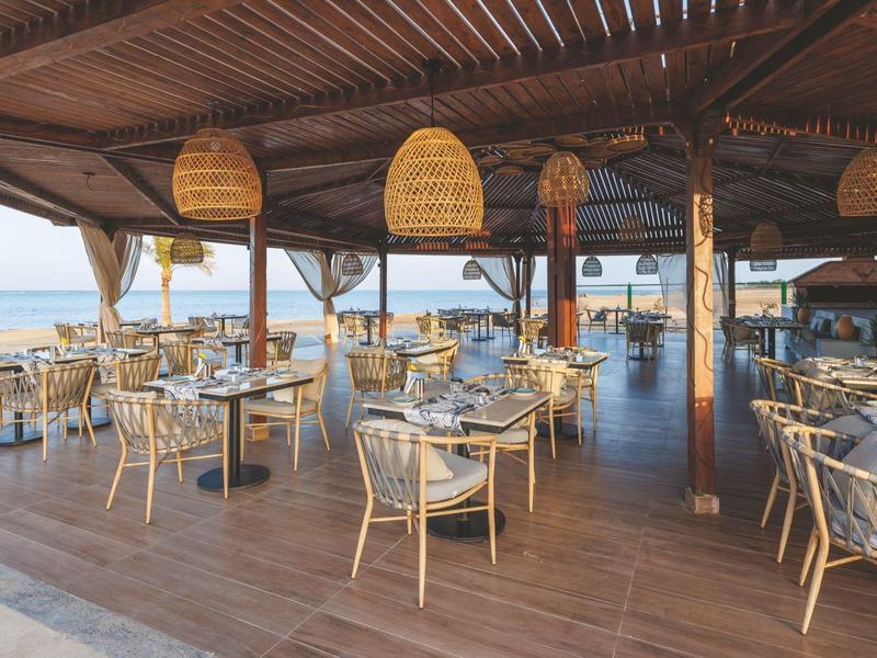 Restaurace na dřevěné otevřené terase s proutěnými židlemi a výhledem na oceán.