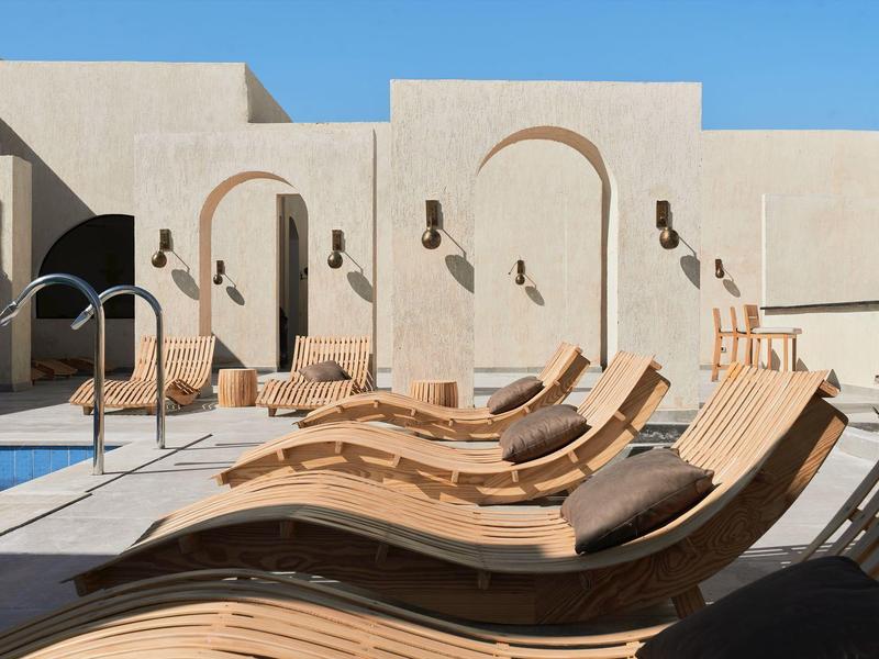 Lettini in legno curvi accanto a una piscina blu sotto un cielo limpido in un resort beige.