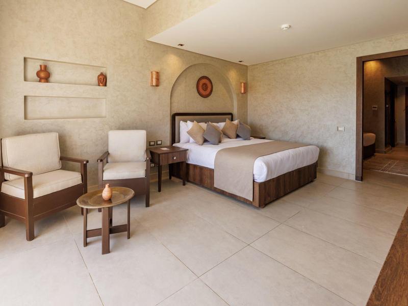 Ampia camera d'hotel con letto grande, due poltrone, tavolino e arredamento neutro.