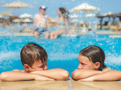 Zwei Kinder lehnen am Beckenrand eines Pools, im Hintergrund sind Menschen und Sonnenschirme unscharf.