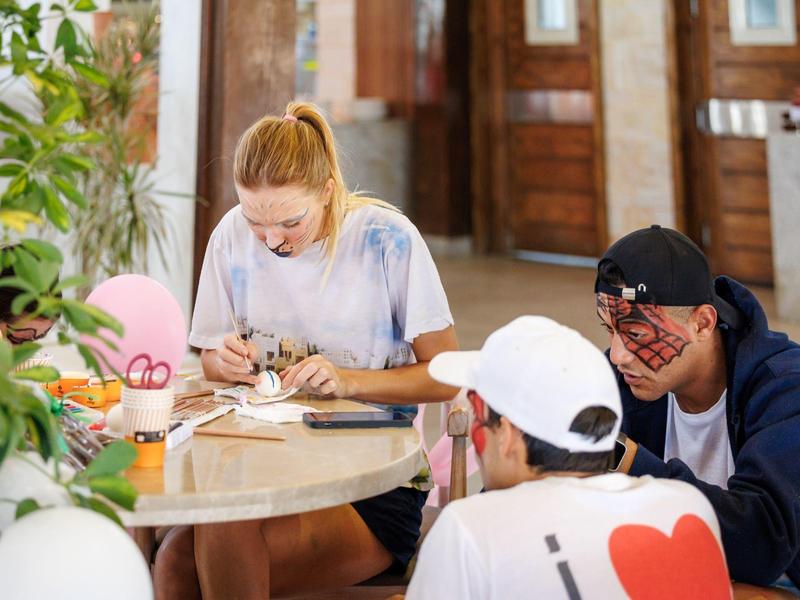 Three young people sitting around a table, engaged in writing or drawing, with balloons nearby.