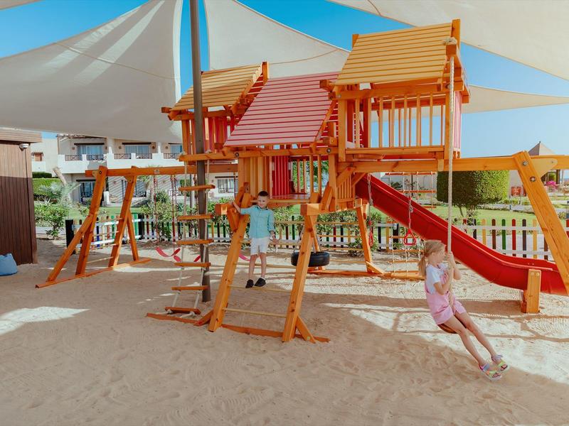 Wooden playground with slides and swings under a sunshade, surrounded by sand.