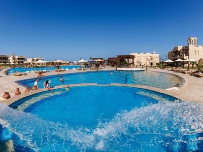 Large hotel pool with clear water, many guests, and surrounding buildings on a sunny day