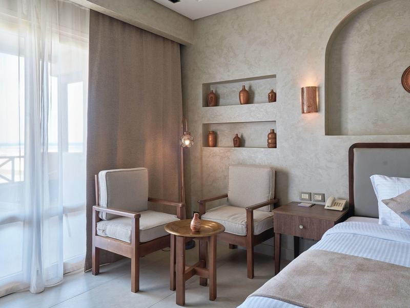 Cozy hotel room with bed, armchairs, small table, soft lighting, and decorative wall shelves.