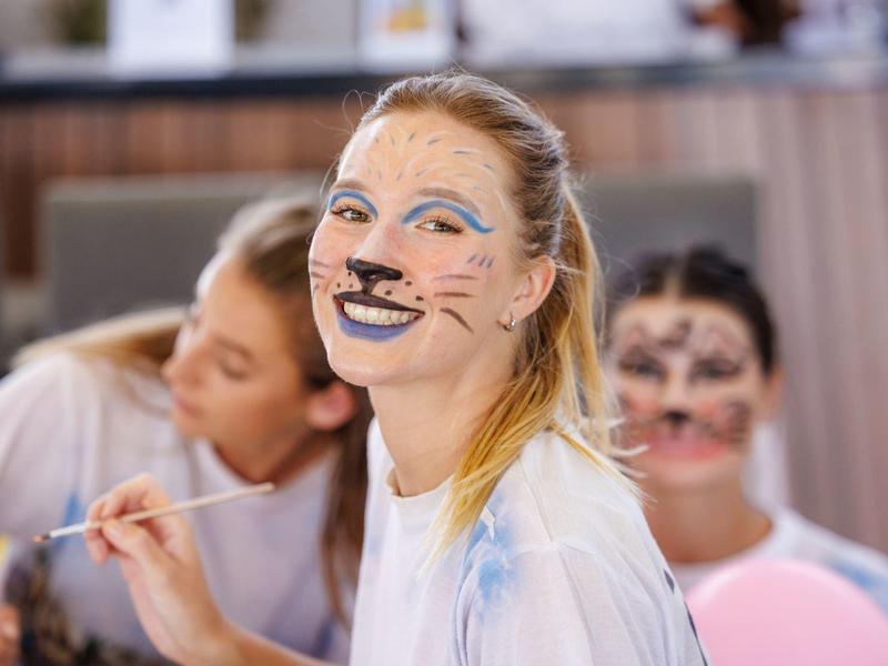 Child with face paint smiling, others in background also painted, indoors, casual setting.