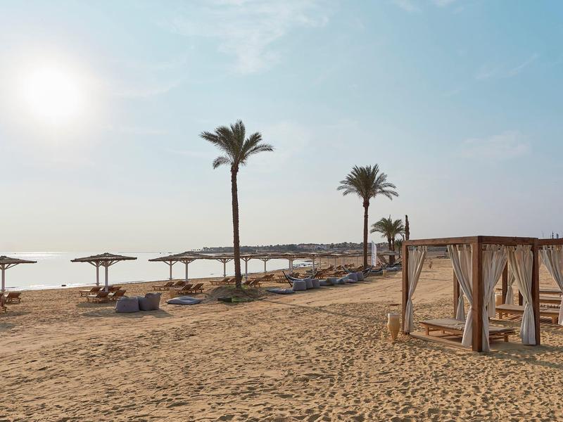 Plage de sable avec chaises longues, parasols, cabanes et palmiers sous un ciel clair.