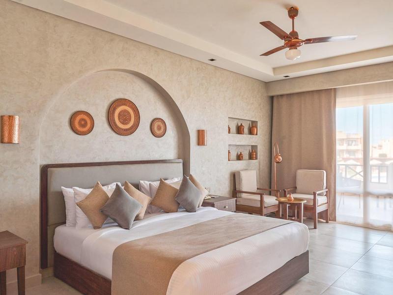 Lichte hotelkamer met groot bed, kussens, zithoek en glazen schuifdeuren naar balkon.