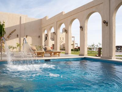 Piscina al aire libre en un hotel con arcos beige y área de descanso bajo el sol.