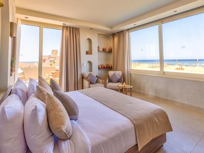 Habitación de hotel luminosa con cama grande, dos sillones y vista al mar a través de grandes ventanas.