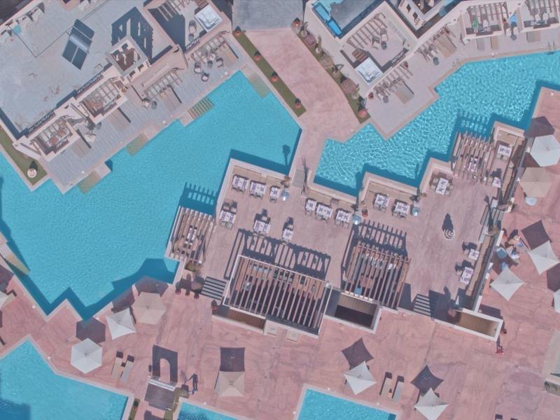 Vista aérea de un hotel con áreas de piscina, sombrillas y tumbonas junto al agua.