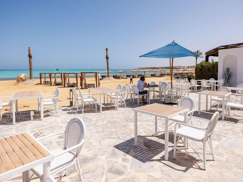 Empty outdoor café seating with white tables and chairs on stone patio by sandy beach and blue sky.