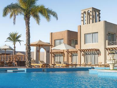 Hotel with pool, palm trees, and sun umbrellas in a sunny setting.