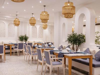 Bright dining area with wooden tables, blue cushioned chairs, hanging lights, and green plants.