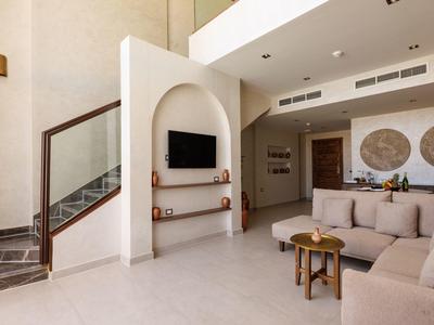 Modern living room with beige sofas, wall-mounted TV, and stone staircase with glass railing.