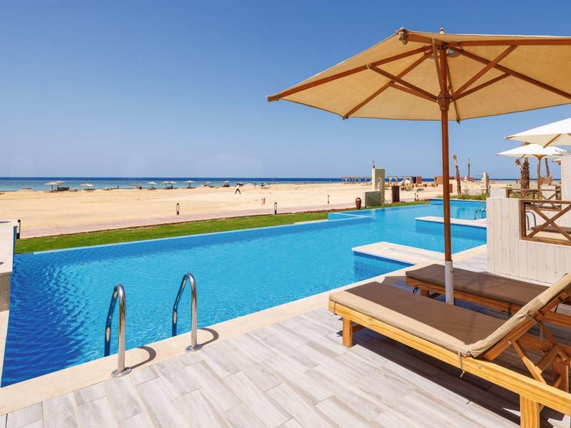 Outdoor pool with sun loungers and umbrellas overlooking a sandy beach and ocean.