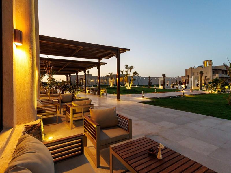Terraza nocturna de un hotel con área de estar, muebles de madera y vista al jardín y edificios.