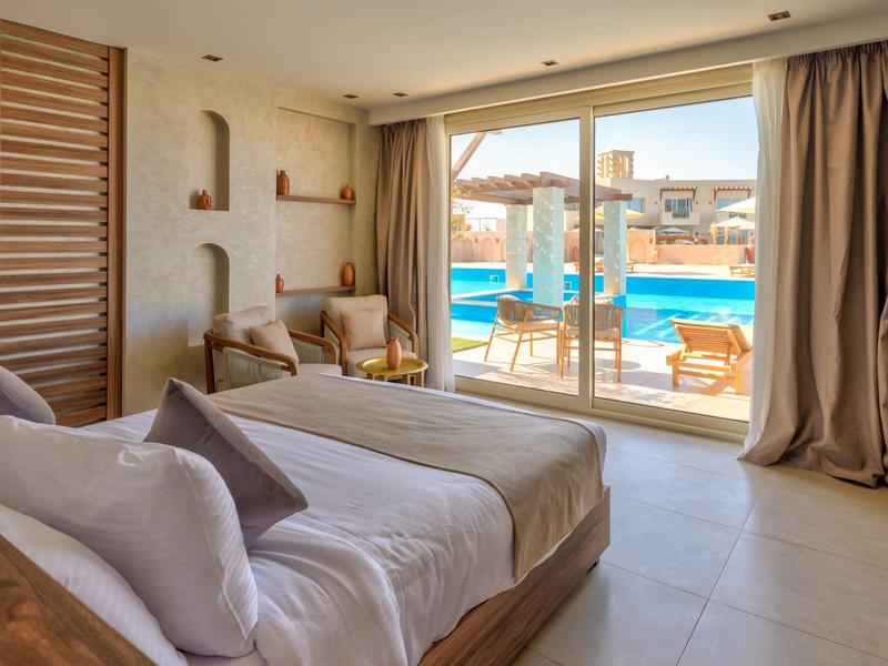 Habitación de hotel luminosa con cama doble y vista a la piscina y sombrillas a través de grandes ventanas.