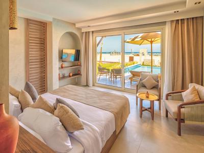 Helles Hotelzimmer mit Doppelbett, Sitzbereich und Blick auf Terrasse mit Pool und Meer.